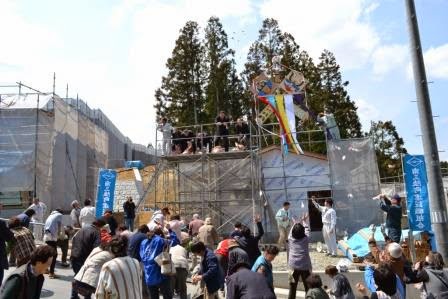 足場の上から餅まきをしている人々と、それを受け取ろうと手を伸ばしている大勢の住民の活気ある上棟式の写真