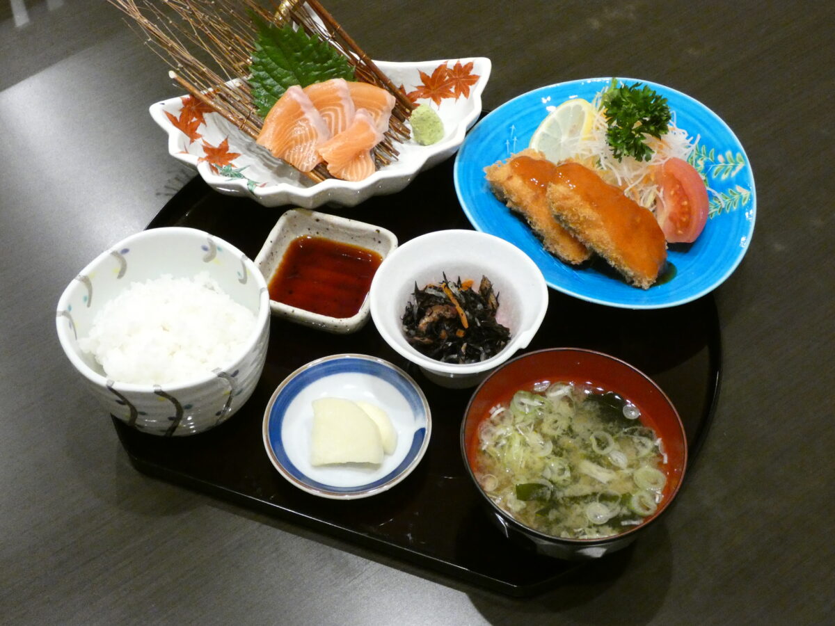 黒いお盆の上に、刺身やフライなどのサケ料理を中心に、白ご飯・味噌汁・漬物・ひじきの煮物・醤油が並べられた料理の写真