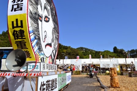 大きな顔のイラストが描かれた山車の横にスピーカーが設置され、背景にはテントが並んだ屋台や人々のにぎわいが見える写真