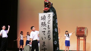 獅子の大きく開いた口が「子どもたち頑張っています」と書かれた紙を咥えて掲げ、背景には存在感のある大太鼓が設置されている舞台上で、白いシャツと青い短パン姿の子どもたちが郷土芸能の発表を行っている写真