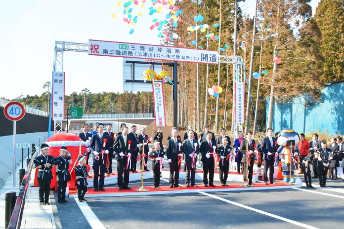 開通を祝う横断幕が取り付けられたゲートの前で、法被姿の子どもたちや関係者が周りに集まる中、胸にリボンをつけた関係者が紅白のリボンを持って整列し、カラフルな風船が空に舞っているテープカットの記念写真