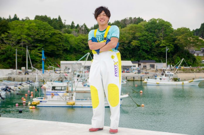 山の手前の複数の漁船が停泊している港を背景に、白と黄色の配色のズボンをはき、腕組みをして笑顔で立っている浅野さんの写真