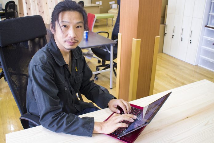 黒い服を着た男性が、オフィスの机に座り赤いノートパソコンを操作しながらカメラを見ている写真