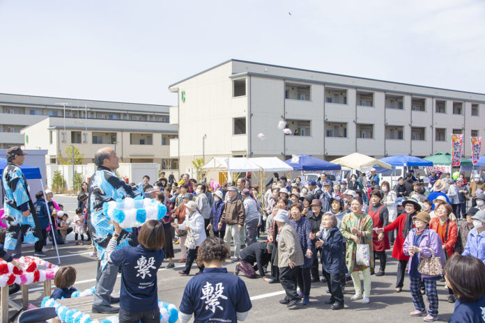 多くの人が屋外で集まり、男性が壇上から紙で包んだ餅を投げ、それを高齢者たちが笑顔で受け取ろうとしている餅まきの様子の写真