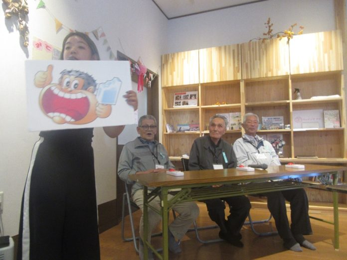 女性が手にマイクを持ち、ガリガリ君のイラストが描かれたパネルを高く掲げて説明しており、前方のテーブルには高齢の男性三人が座って話を聞いているクイズ大会の様子の写真