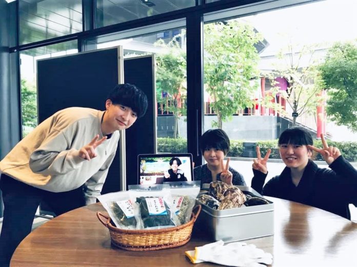 3人の大学生が室内の丸いテーブルの前でピースサインをして笑顔で写っており、テーブルには乾燥海藻の袋と缶に入った牡蠣が置かれ、中央にはオンライン通話中の人物が写るタブレットがある写真