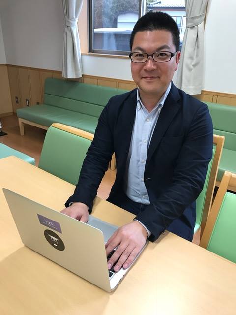 窓際に若草色の座面の長椅子が置かれた部屋で、テーブルに置いたノートパソコンのキーボードに両手を置いて座っている佐藤さんの写真