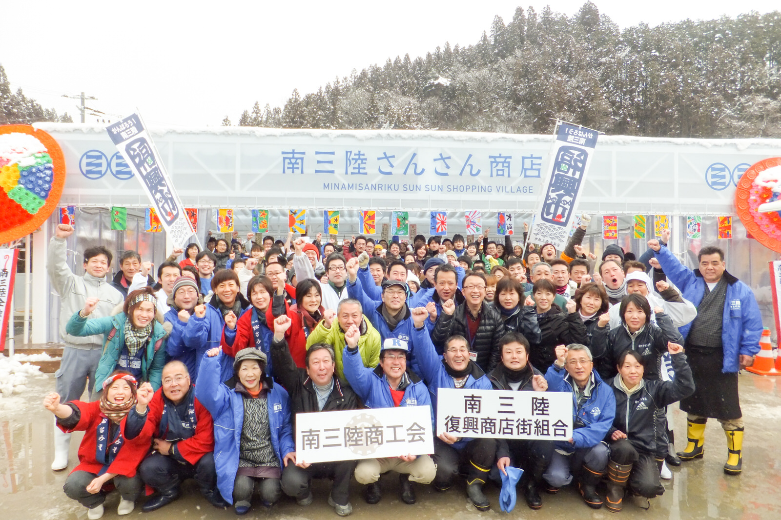 雪が降る中、「南三陸商工会」「南三陸復興商店街組合」の看板を掲げている沢山の人々が「南三陸さんさん商店街」と書かれた建物の前で笑顔で拳を上げて並んでいる集合写真