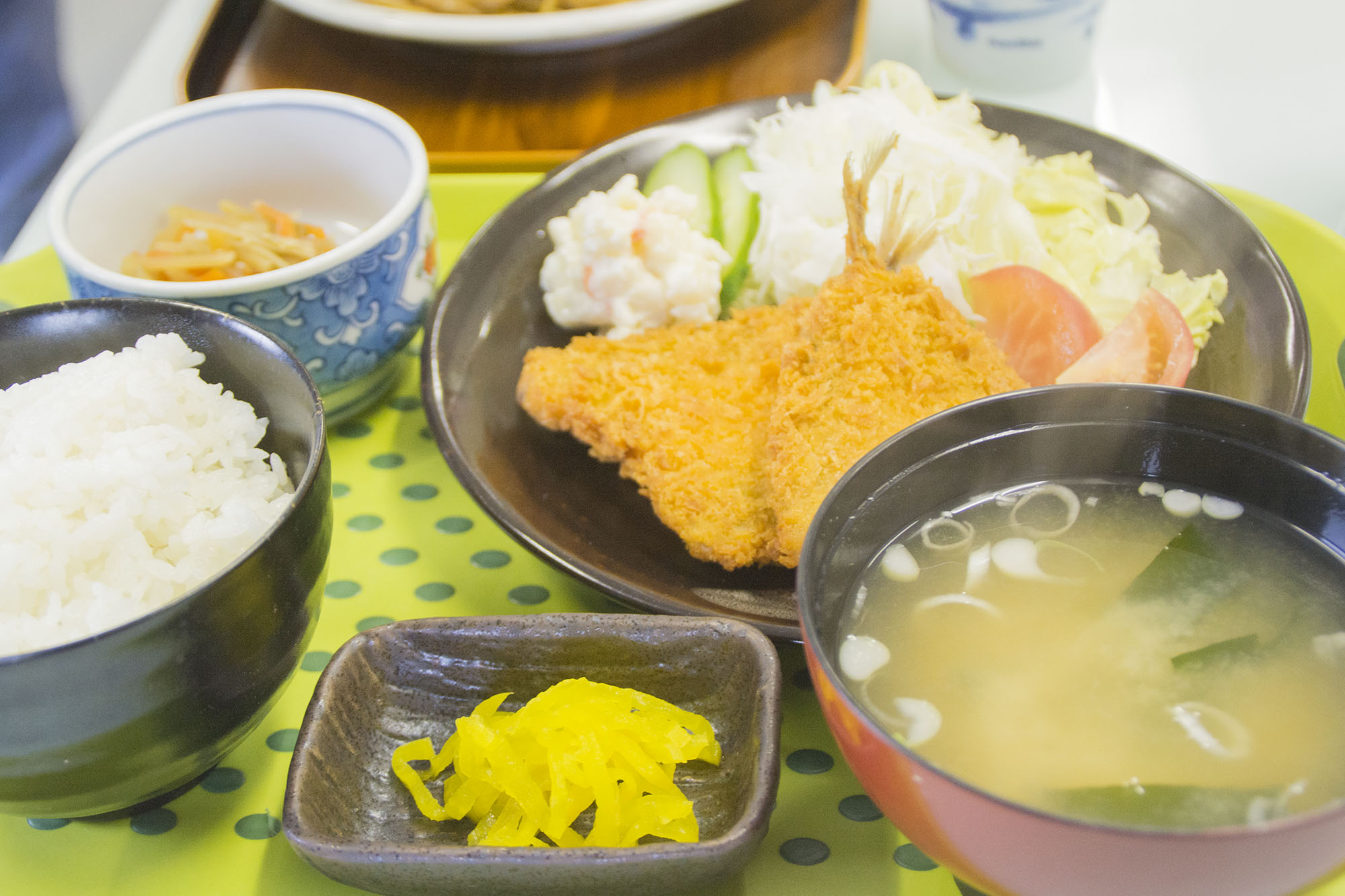 緑のトレイの上に、ご飯、味噌汁、漬物、小鉢、ポテトサラダと千切りキャベツを添えたアジフライが盛られた料理が並んでいる定食のアップ写真