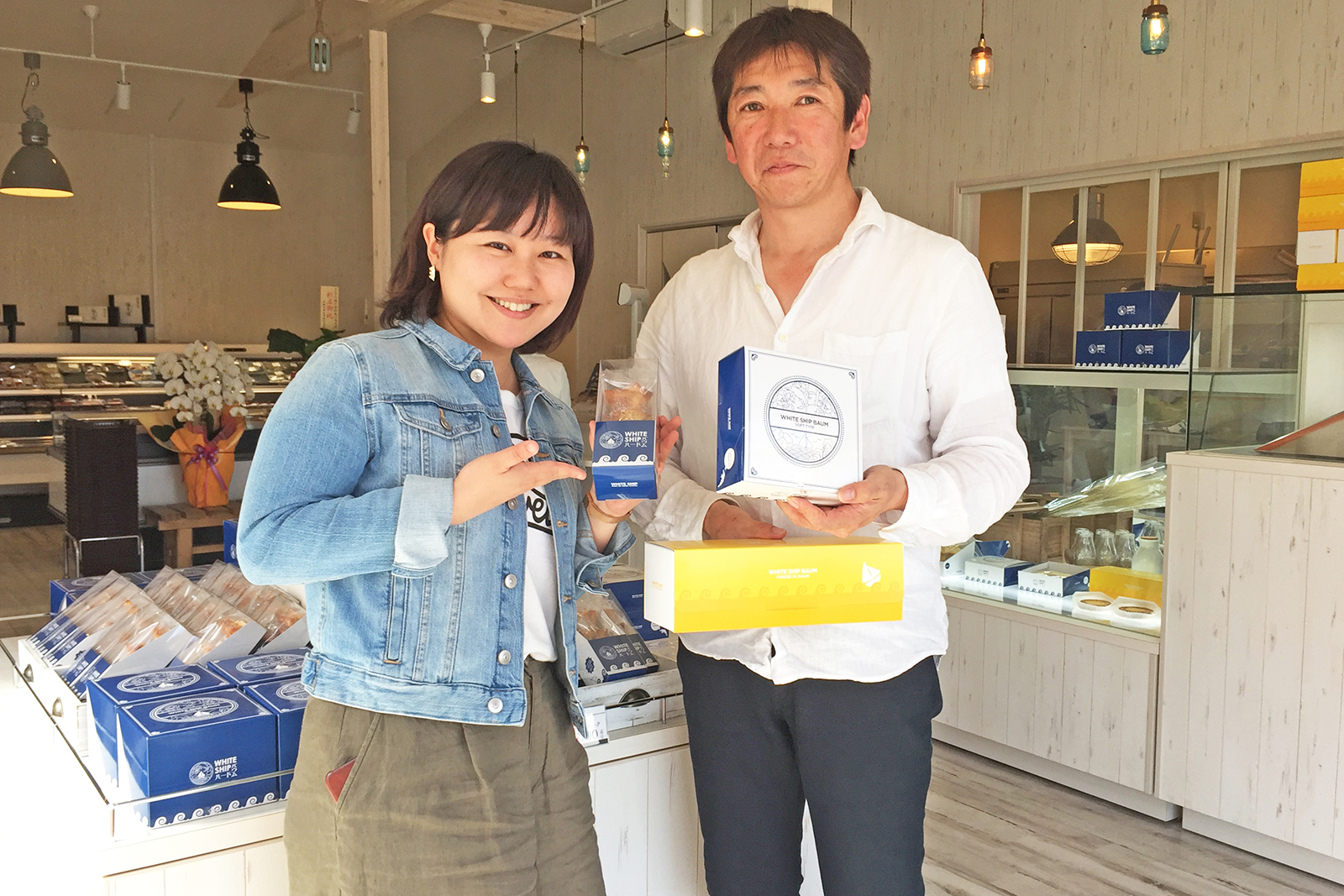 陳列棚やショーケースにいろいろなバームクーヘンの商品が並んだおしゃれな店内で商品を手に並んで立った女性リポーターと男性の写真