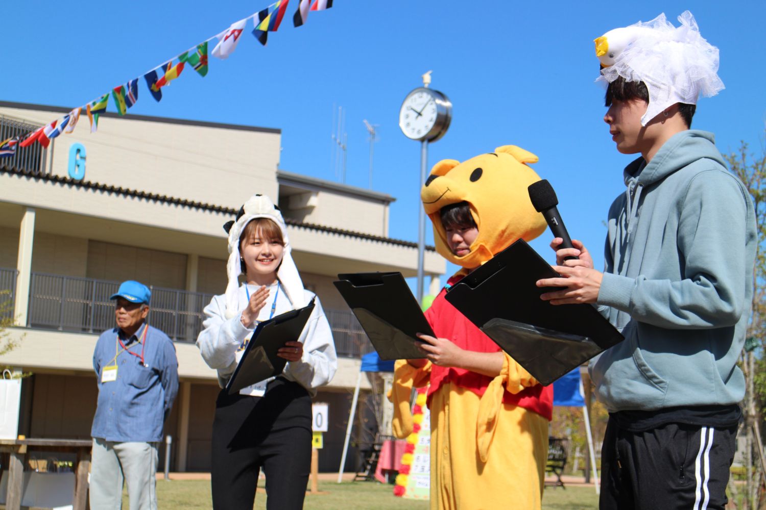 青空の下、動物の着ぐるみや帽子をかぶっている3人がマイクを持ち、司会進行する様子の写真