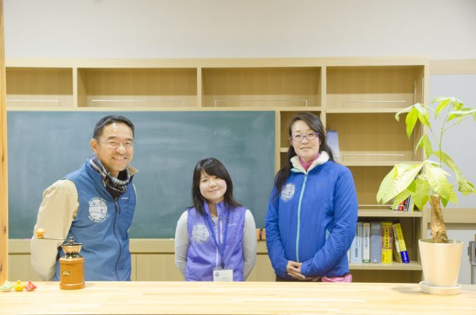 本棚と黒板がある室内で、お揃いで色違いのユニフォームを着た三人のスタッフがカウンターの前に並び笑顔で写っている写真