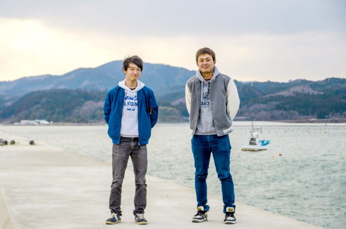 海辺の堤防で山並みを背景に2人の男性が並んで立っている写真