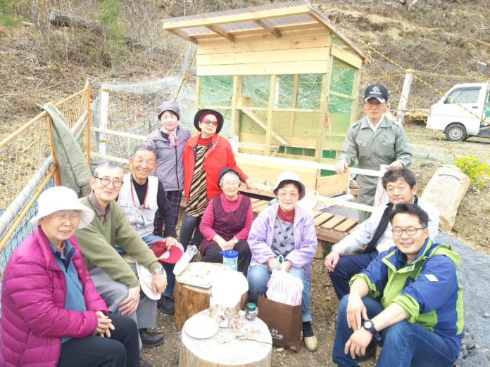 木製の小屋の前で、色とりどりの服を着た十人ほどの地域の方々が丸太の椅子に座っている集合写真