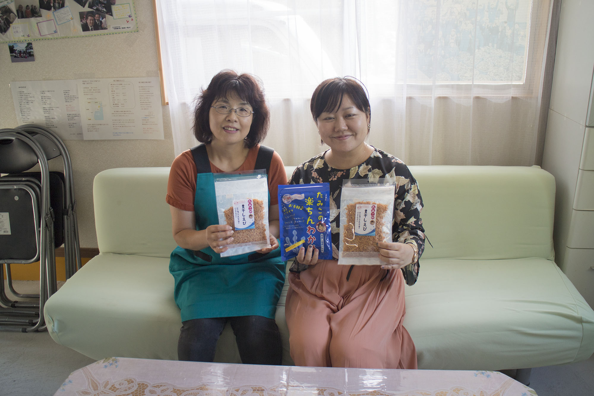 室内のソファに座ったエプロン姿の阿部民子さんと女性リポーターが、パックに入った商品を手に持って笑顔でカメラを見ている写真