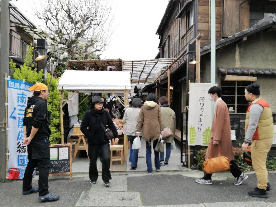 和風の建物が並ぶ通りの一角にある「三陸ワカメまつり」と書かれた看板が設置された会場入り口で、何人かの来場者が白いテントの脇の通りに入っていく様子が写されている様子の写真
