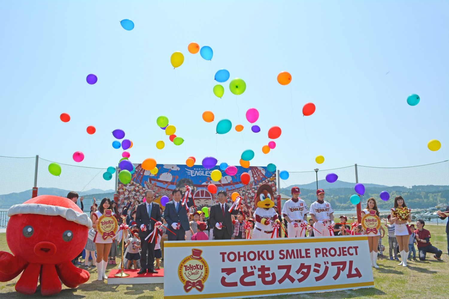 「TOHOKU SMILE PROJECT こどもスタジアム」と書かれたプレートの前で、南三陸町のマスコットキャラクターオクトパス君と楽天イーグルスのキャラクターや選手、子どもたち、関係者が集まり、カラフルな風船を空に飛ばしているテープカットが行われた様子の写真