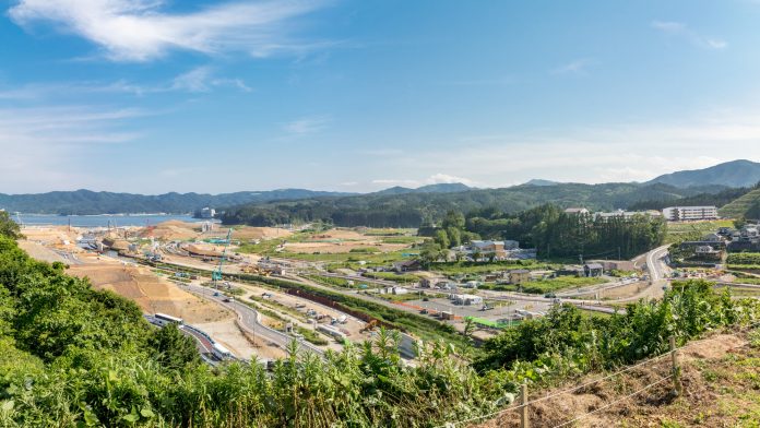 造成が進んだ海沿いの町の定点カメラによる風景で抜けるような青空の中左奥には穏やかな海が広がっており、防波堤やクレーン船がいくつか確認できその手前では大規模な土地造成が進められており、土を盛って造成された大きな段差（盛土）が連なって大型のクレーン車やショベルカーが稼働している現場の写真