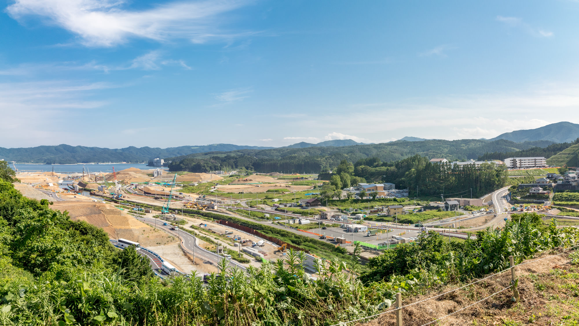 快晴の中、復興造成が進む海辺の町の定点カメラによる風景で左端には海が広がっており、沿岸には多くの重機や資材が集まり造成中の土山や重機（ショベルカー、クレーンなど）が並んでいる、大規模な土木工事の様子を写した写真