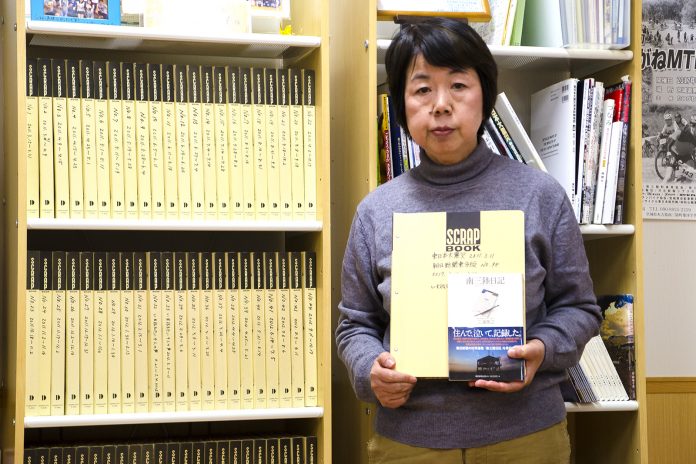 薄い黄色の背表紙のスクラップブックがずらりと並んだ本棚の前で、スクラップブックと本を胸の前で抱えた越後圭子さんが立っている写真