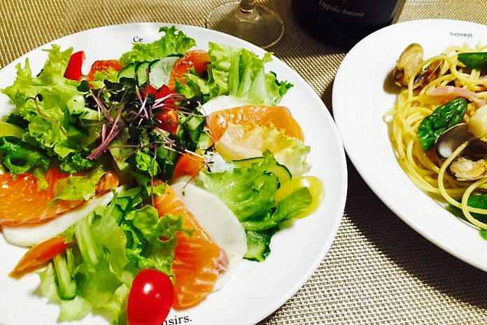 新鮮なサーモンの刺身が数枚、薄くスライスされ白い皿の上に盛り付けられていて、その周りにレタス、きゅうり、トマト、赤パプリカ、カイワレなどの色鮮やかな野菜が彩りよく盛り付けられている、食卓のサラダとパスタの料理の写真