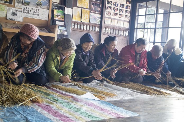 地区の高齢者の皆さんが部屋の中で一列に座り、稲わらを手に楽しく会話しながら縄をなう作業に取り組んでいる様子の写真