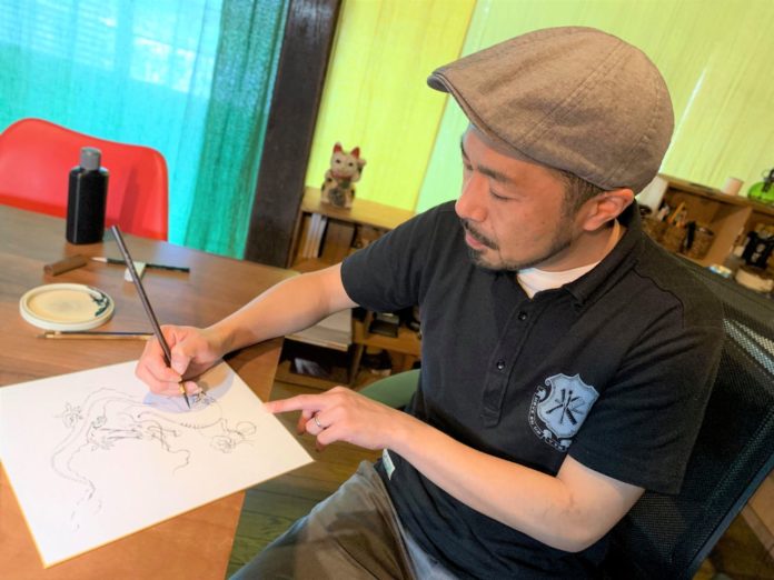 机に黒いボトルと筆皿が置かれている部屋で、グレーのキャスケット帽をかぶった男性が木製の机に向かい、筆ペンを使って白い紙に龍の線画を描いている様子の写真