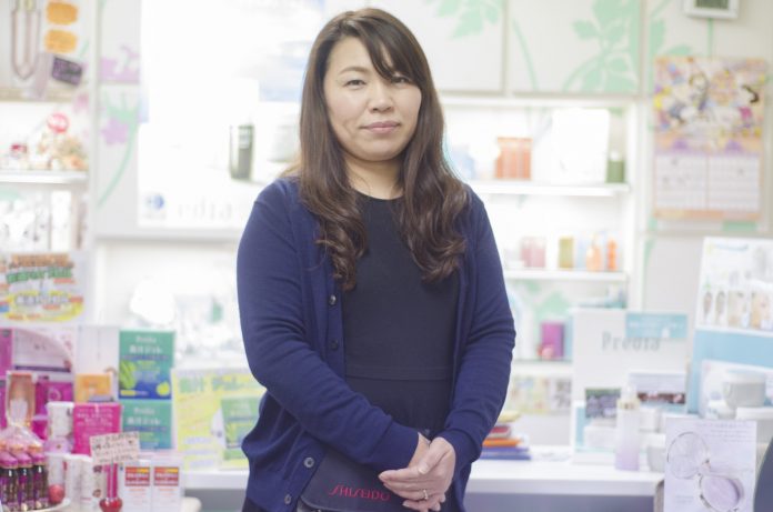 化粧品棚の前に立つ、両手を前で軽く組んで正面を見つめている薄紫色のカーディガンを着た女性をアップで撮影した写真