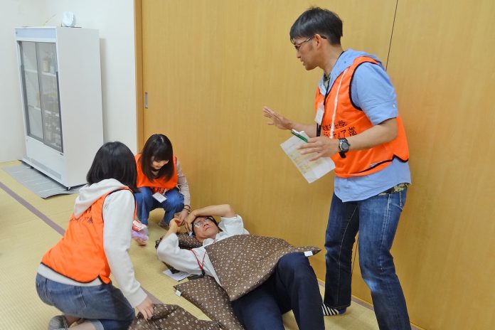 防災訓練中に、床に倒れている男性に対してオレンジ色のベストを着た3人の大学生が応急手当の練習を行い、もう1人の男性が立って指示を出している様子の写真