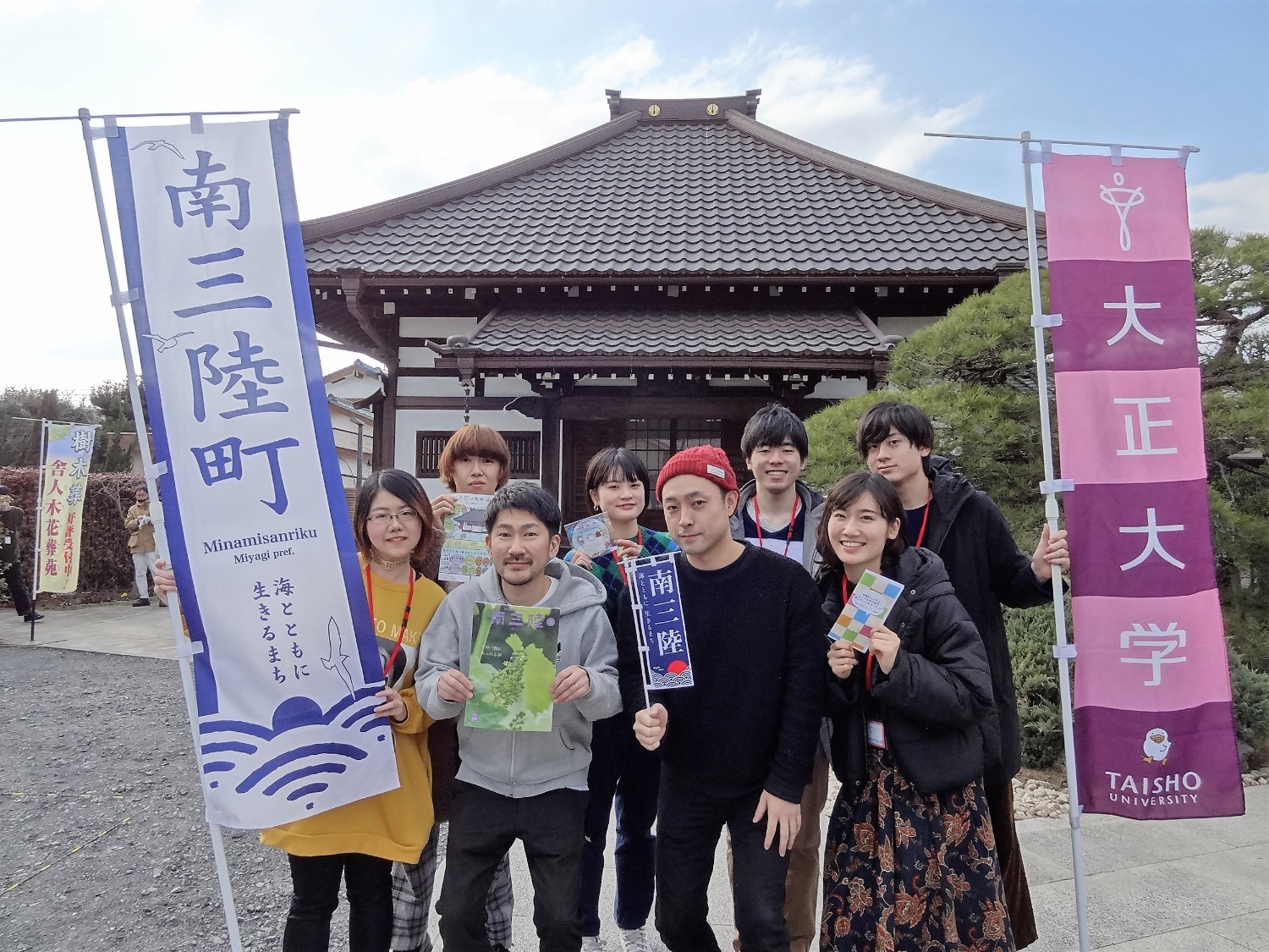 お寺の前で「南三陸町」と「大正大学」ののぼりを持った若者たちが、笑顔で記念撮影をしている写真