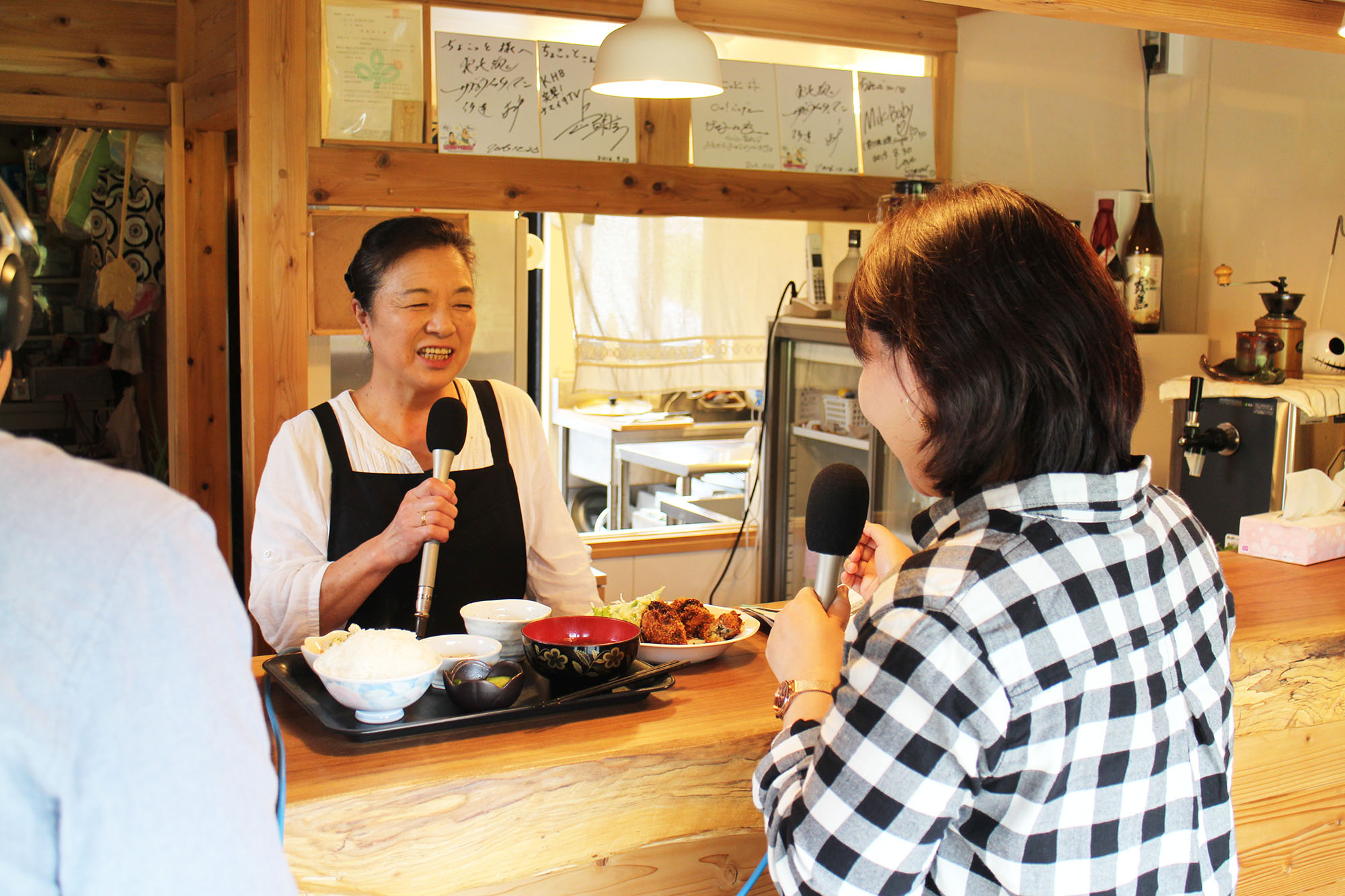 飲食店のカウンター越しに、カキフライ定食を前にしてマイクを持ちながら笑顔で話す成澤さんと、インタビューをしている女性リポーターの写真