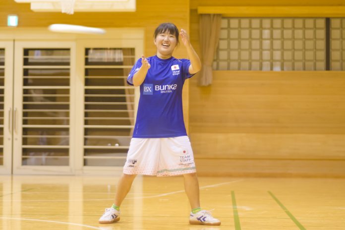 体育館で青いシャツと白いショートパンツを着てポーズをとっている様子の笑顔の宮岡茜さんの写真