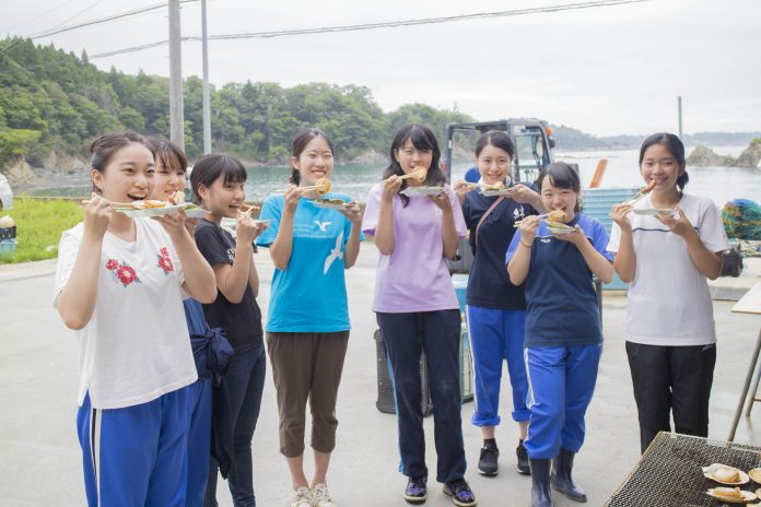 恵泉女学園高等学校OG8人が海を背景に並び、焼いた貝を箸で頬張りながら笑顔を見せている写真