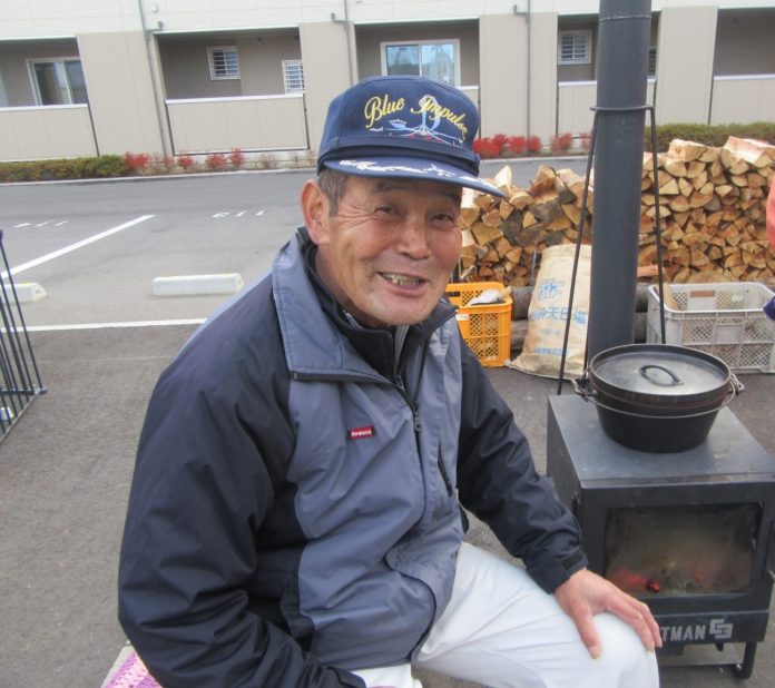 帽子をかぶって屋外の薪ストーブの前に座り笑顔を見せている佐藤良夫さんの写真