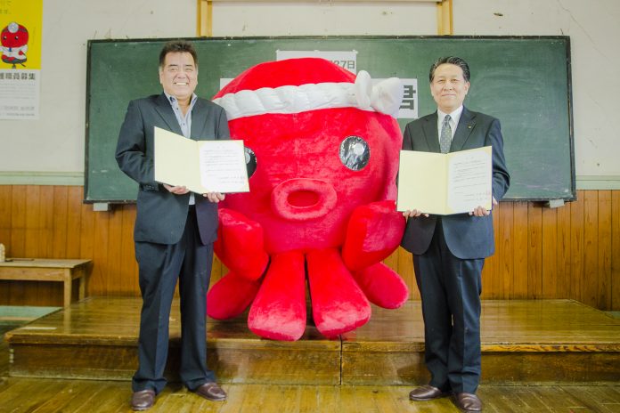 黒板の前に立ったオクトパス君の着ぐるみの前に並んだ佐藤さんと高橋さんが調印した文書を胸の前で持って笑顔で写っている記念写真