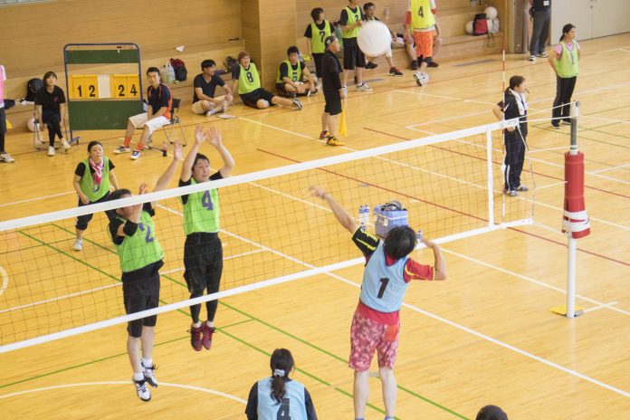 体育館の中で、緑と青のビブスを着た地域住民が混成チームでバレーボールをプレイしており、観客が周囲に座って試合を見守っている写真