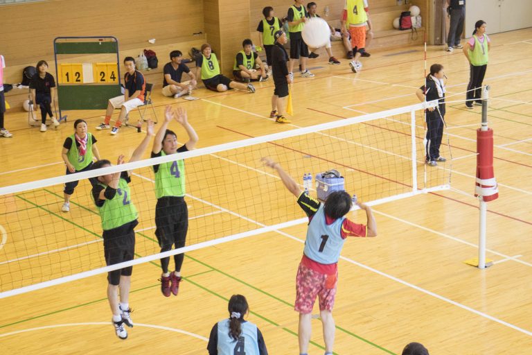 体育館の中で、緑と青のビブスを着た地域住民が混成チームでバレーボールをプレイしており、観客が周囲に座って試合を見守っている写真
