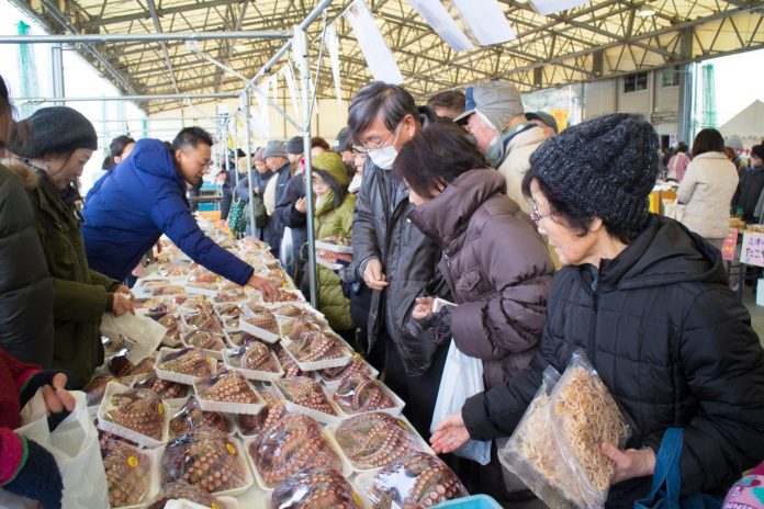 福興市の出店で、多くの人がテーブルに並べられた多数のタコ足のパックを見ながら、店主と会話をしたり、商品を手に取って選んでいる様子を捉えた写真