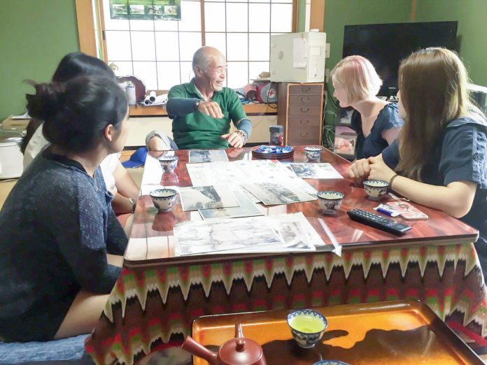 畳の部屋で、コタツで暖まりながら年配の男性が若い4人の女性たちに昔の写真を見せながら楽しそうに語り合っている様子の写真