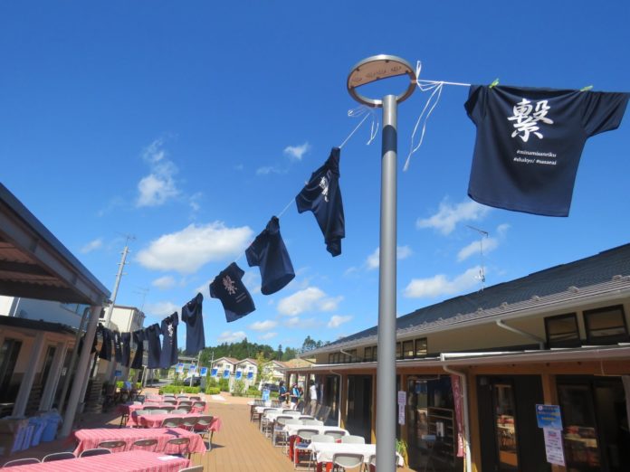 青空の下、屋外のウッドデッキに赤いテーブルクロスがかけられた多数のテーブルと椅子が並べられ、その上には紐に吊るされた紺色のTシャツが風になびき、右側のTシャツには「繋」という漢字が白字で書かれている写真