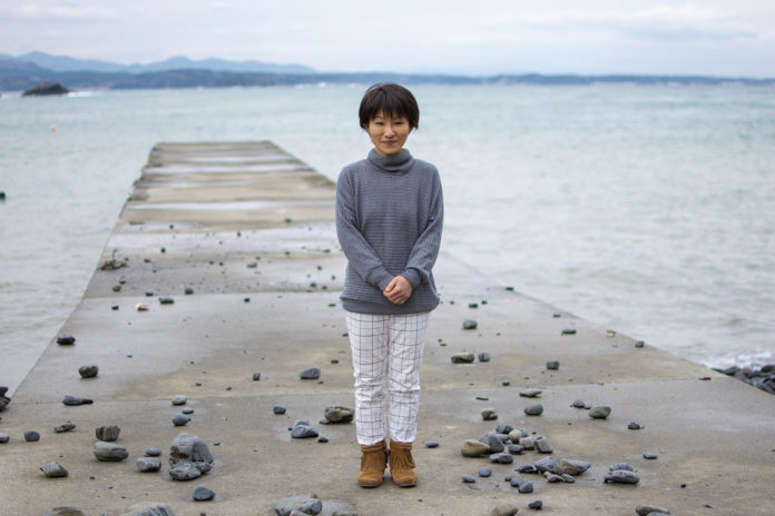 海に突き出したコンクリートの桟橋の上で、灰色のタートルネックと白いチェックのズボンを履いた女性が正面を向いて微笑んでいる写真