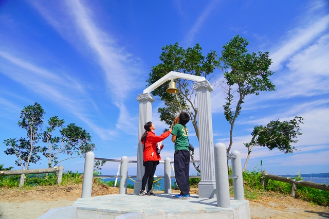 青い空にうっすらとした白い雲が伸びている晴れの日に、古代ギリシャ風のコンクリート製の柱に取り付けられた幸せの鐘を鳴らす男女の写真
