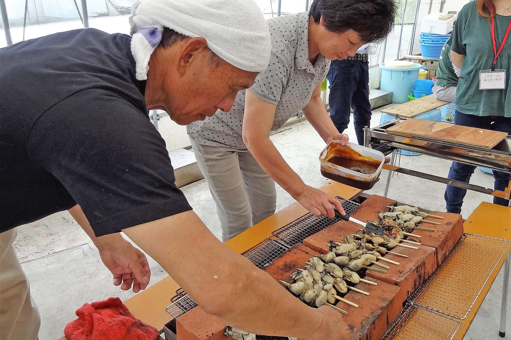 漁師さんと女性が串に刺したカキを炭火で焼きながらタレを塗っている写真
