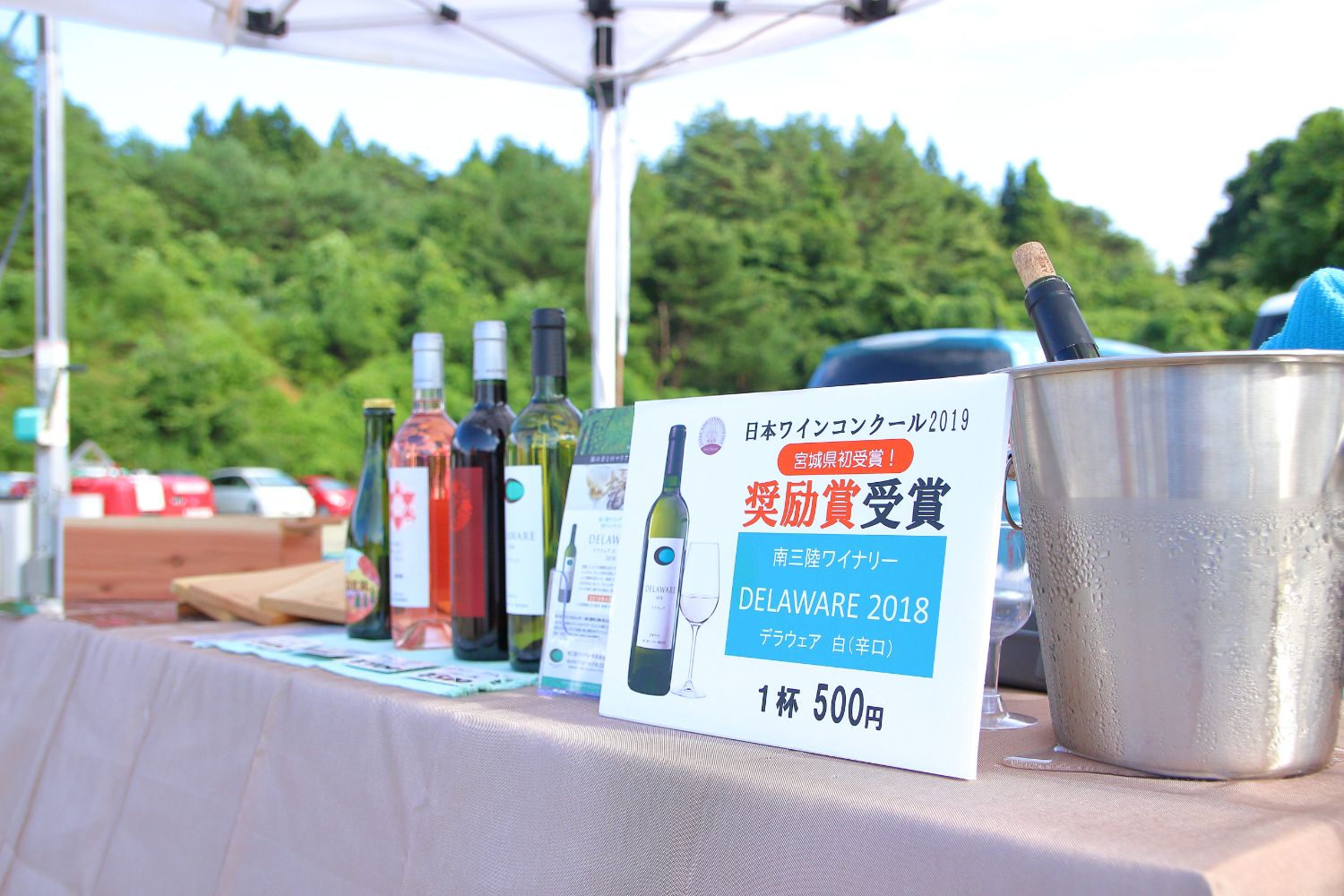 「日本ワインコンクール2019奨励賞受賞」と書かれた南三陸ワイナリーの紹介パネルが置かれているテントの下に、ワインボトルが並んでいる写真