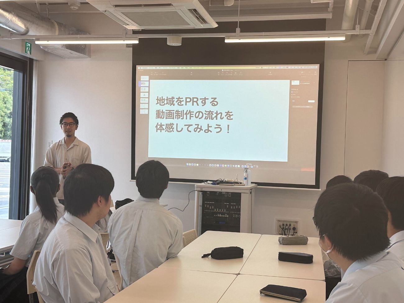 スクリーンには「地域をPRする動画制作の流れを体感してみよう！」というスライドが映し出され男性の話を真剣に聞いている高校生たちの写真