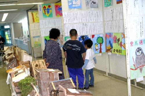 屋内の展示スペースで、大人1人と子ども2人が壁に貼られた児童の描いた鳥の絵や標語のようなもの、そして床に並べられた手作りの鳥の巣箱を鑑賞している、愛鳥週間作品展の様子の写真