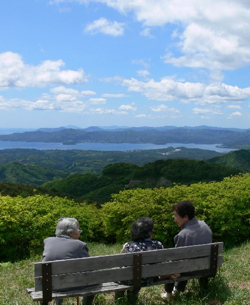 広大な緑の山々と、その先に青い海と空が広がる絶景を背景に、3人の年配の人物がベンチに座り、遠くの景色を眺めながら会話している様子の写真