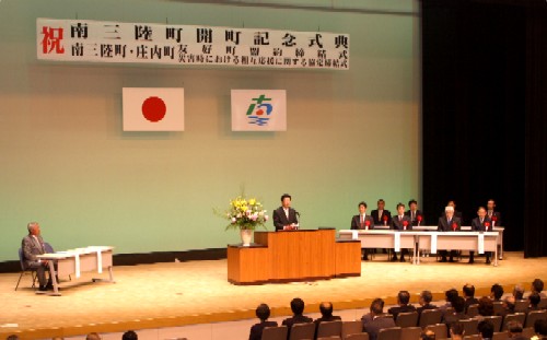 照明が当たった広いステージで、スーツ姿の男性が中央の演台でスピーチをしており、後方の横長のテーブルには来賓らしき関係者が並んで座っている開町式の様子の写真