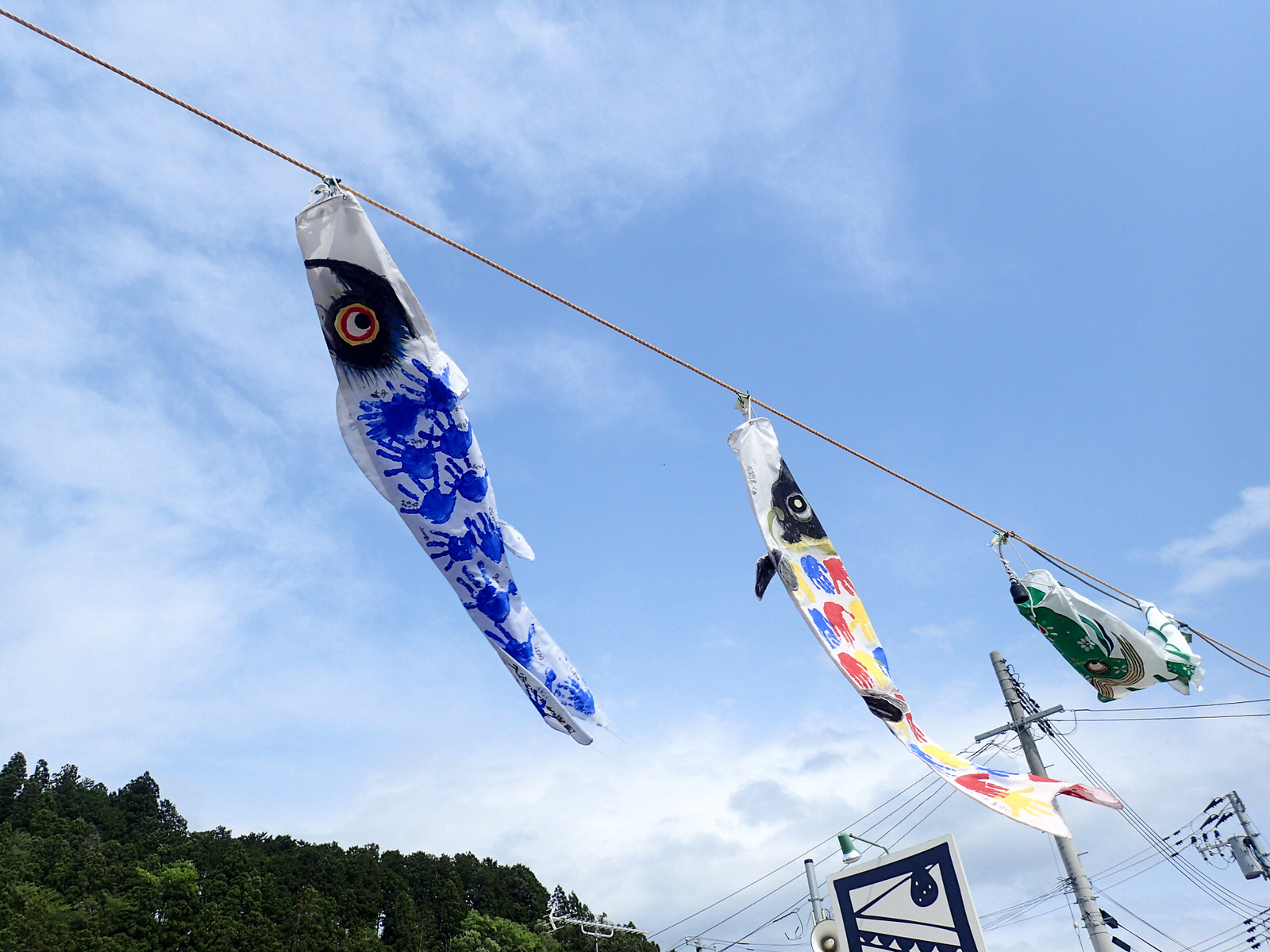 青空の下、子どもの手形で彩られた青・黄・緑の鯉のぼりがロープに吊るされて風に揺れている様子の写真
