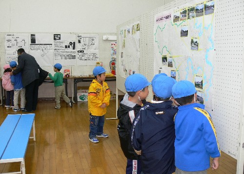 青い帽子をかぶった数人の子どもたちが、壁に貼られた大きな地図や写真を熱心に眺めており、左奥では関係者と他の子どもたちが話をしている津波資料展の様子の写真
